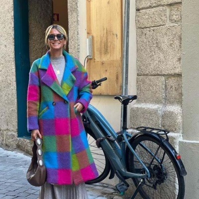 Mulher com casaco xadrez colorido numa rua com bicicleta elétrica ao lado.