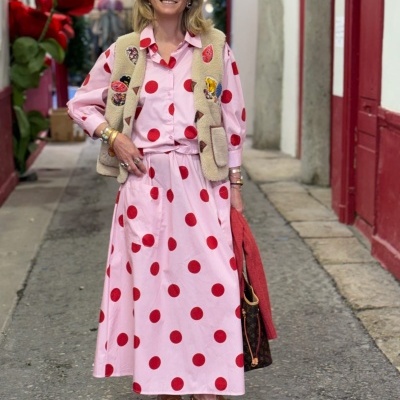 Mulher com vestido rosa de bolinhas vermelhas, colete bege felpudo e chapéu de palha numa rua urbana