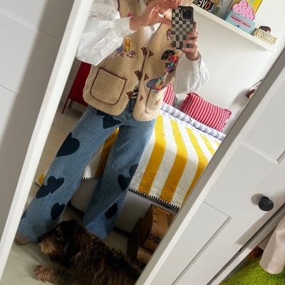 Mulher com colete de pelo bege com patches e calças de ganga com corações a tirar selfie num espelho