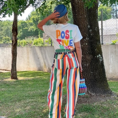 Pessoa de costas com camiseta branca com texto colorido e calças riscas verticais coloridas em ambiente exterior