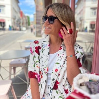 Mulher com casaco e calças florais, óculos de sol e anel vermelho numa esplanada