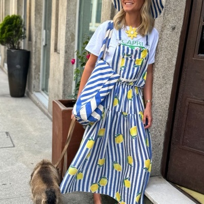 Mulher com vestido riscado azul e branco com limões, chapéu e bolsa às riscas, t-shirt branca CAPRI, sapatos verdes na rua com cão