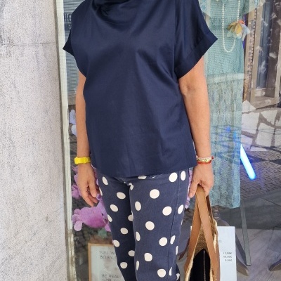 Pessoa vestindo blusa azul escura e calças azul marinho com bolinhas brancas, sandálias castanhas, com cabelo branco e óculos escuros.