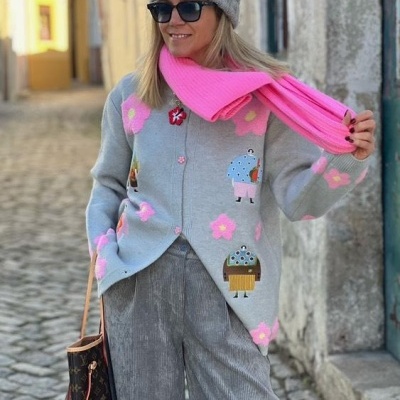 Mulher com casaco cinza de malha com botões rosa e padrão de flores coloridas, calças cinza, cachecol rosa, gorro cinza e óculos escuros