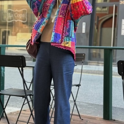 Mulher veste casaco patchwork colorido, calças azuis largas, camisola bege, meias rosas, mocassins beges, boina branca e óculos escuros.