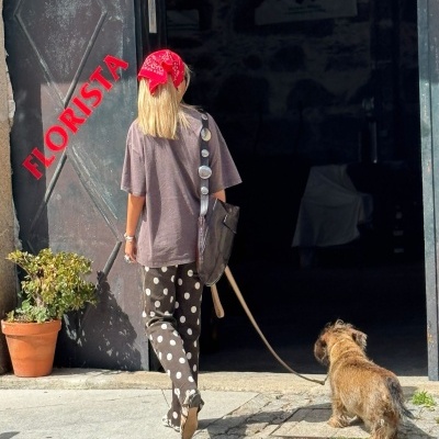 Mulher com bandana vermelha e calças de bolinhas entrando em loja com porta preta e texto FLORISTA, acompanhada de cachorro pequeno.