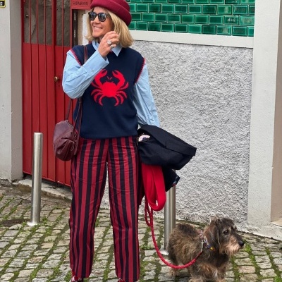 Mulher com colete azul e caranguejo vermelho, calças riscas vermelhas e azul, com cão pequeno numa trela vermelha numa rua com calçada de pedra.