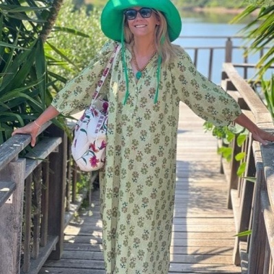 Mulher com vestido comprido verde claro com flores, chapéu verde, óculos de sol e saco floral num passadiço de madeira perto de um rio.