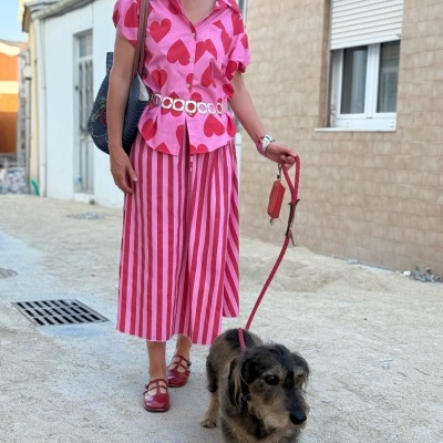 Mulher com blusa rosa de corações vermelhos e calças às riscas rosa e vermelhas a passear cão com trela vermelha na rua