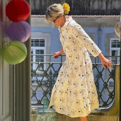 Vestido midi amarelo claro com padrão de personagens, usado por mulher junto a varanda urbana