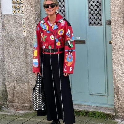 Pessoa com casaco vermelho pintado, calças pretas, bolsa preta com bolinhas, sapatos com bolinhas, óculos escuros, porta azul clara ao fundo