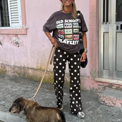 Mulher com t-shirt estampa OLD SCHOOL PLAYLIST e calças com bolinhas, segurando trela de cão castanho numa rua ao lado de muro rosa