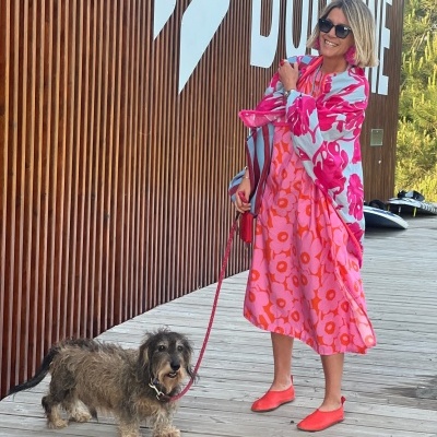 Mulher vestida com vestido rosa de flores e capa azul com flores cor-de-rosa segurando trela de cão peludo.