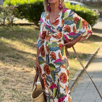 Mulher a usar vestido longo branco com padrão colorido de folhas e flores e a segurar mala de palha.
