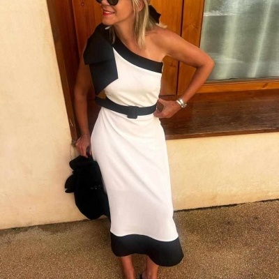 Mulher a usar vestido branco e preto com cinto, óculos escuros e chinelos pretos junto a parede com janela de madeira