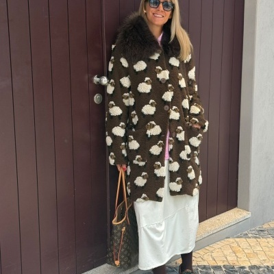 Mulher com casaco castanho de ovelhas, vestido branco, sapatos castanhos, óculos escuros e bolsa monograma em frente a porta castanha.