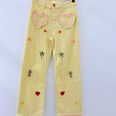 Calças amarelas xadrez com bordados coloridos e bolsos em forma de coração penduradas num cabide