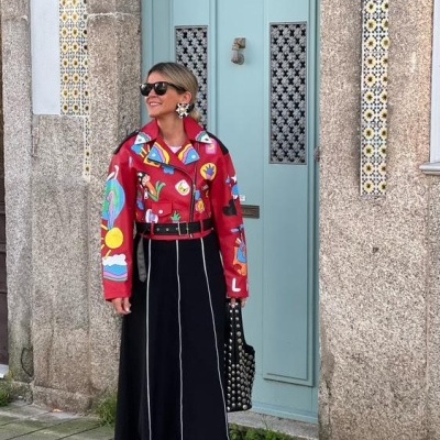 Mulher vestindo casaco vermelho estampado, saia preta longa e sapatos com bolinhas, em frente a porta azul e parede de pedra com azulejos
