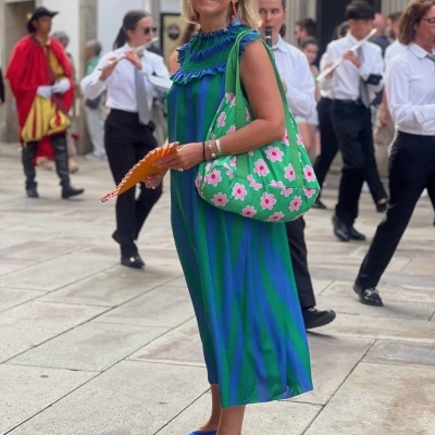 Mulher sorridente com vestido azul e verde, óculos, bolsa com flores, sapatos azuis e leque laranja num espaço urbano com pessoas ao fundo.