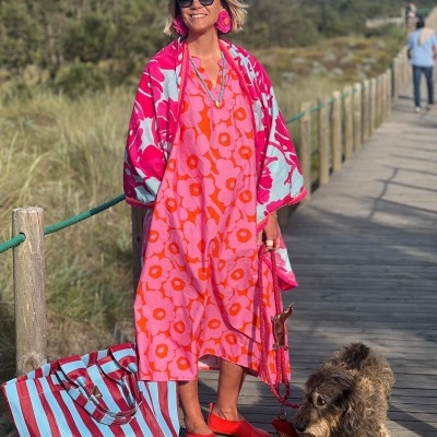Mulher com vestido rosa de flores laranja, manta rosa e azul, brincos florais, sapatilhas vermelhas, mala riscada azul/vermelho, coleira vermelha, com cão castanho preto num passadiço de madeira.