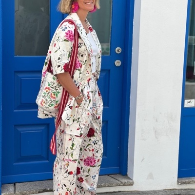 Mulher com conjunto floral branco, bolsa têxtil, sapatos vermelhos e óculos escuros junto a porta azul