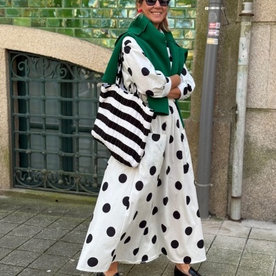 Mulher com vestido branco às bolinhas pretas, carteira às riscas e casaco verde sobre os ombros.
