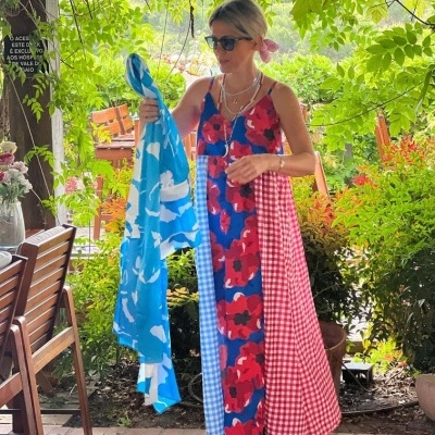 Mulher com vestido longo colorido e padrões variados, segurando tecido azul, em área externa com vegetação e cadeiras de madeira
