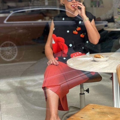 Mulher sentada ao lado de mesa redonda num café, vestindo camisola e saia vermelhas e sapatos pretos decorados.