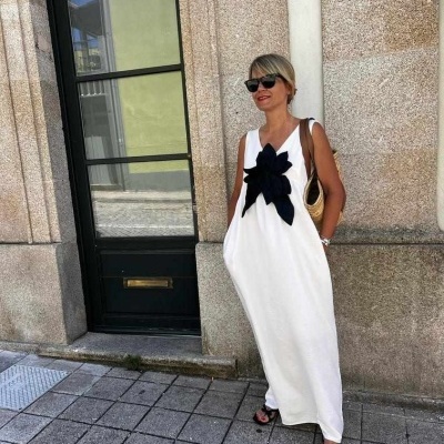 Mulher vestida com vestido comprido branco com flor preta, óculos de sol, sandálias pretas, junto a prédio urbano.