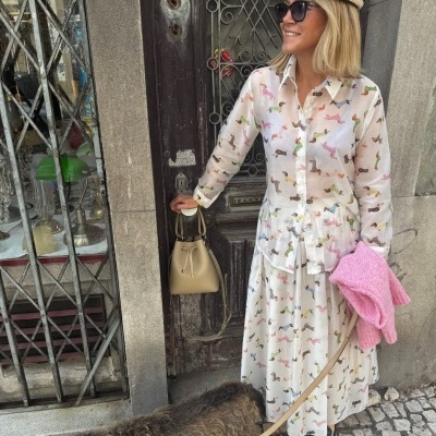 Mulher com vestido branco estampado de cães e acessórios, em frente a porta de madeira com cão ao lado.