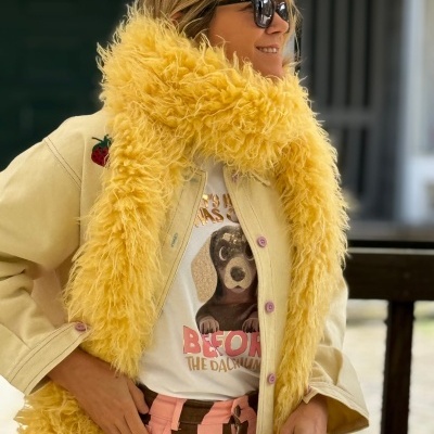 Pessoa com lenço amarelo felpudo, camisola com cão e texto, casaco bege com bordado e calças riscas castanho e rosa