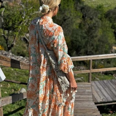 Mulher com vestido floral comprido e mala de crochê cinza num passadiço de madeira ao ar livre
