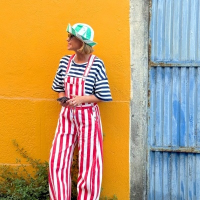 Pessoa com roupa colorida a pé encostada em parede amarela junto a porta azul metálica