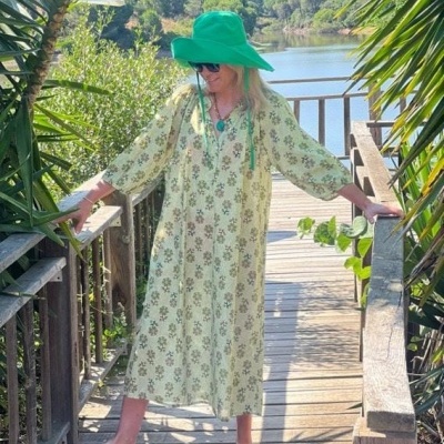 Mulher com vestido floral verde e bege, chapéu verde e chinelos em passadiço de madeira perto de lago