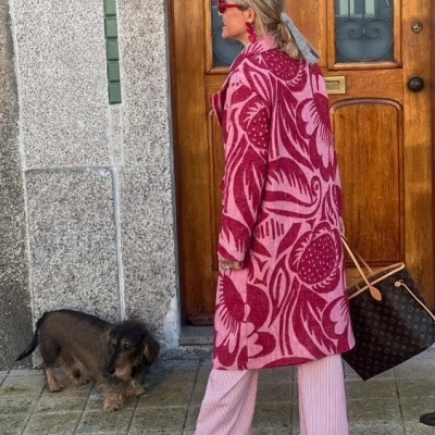 Mulher com casaco rosa e vermelho e ténis vermelhos segura bolsa castanha à porta de madeira junto a cão