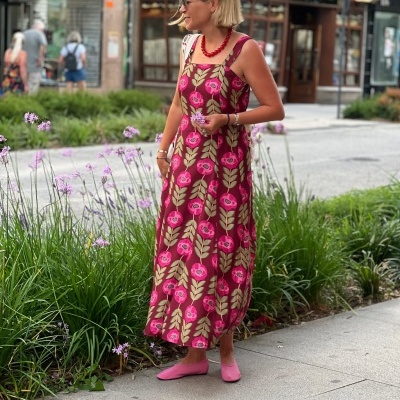 Mulher com vestido floral rosa e verde, bailarinas cor de rosa e colar vermelho na rua perto de plantas com flores lilás