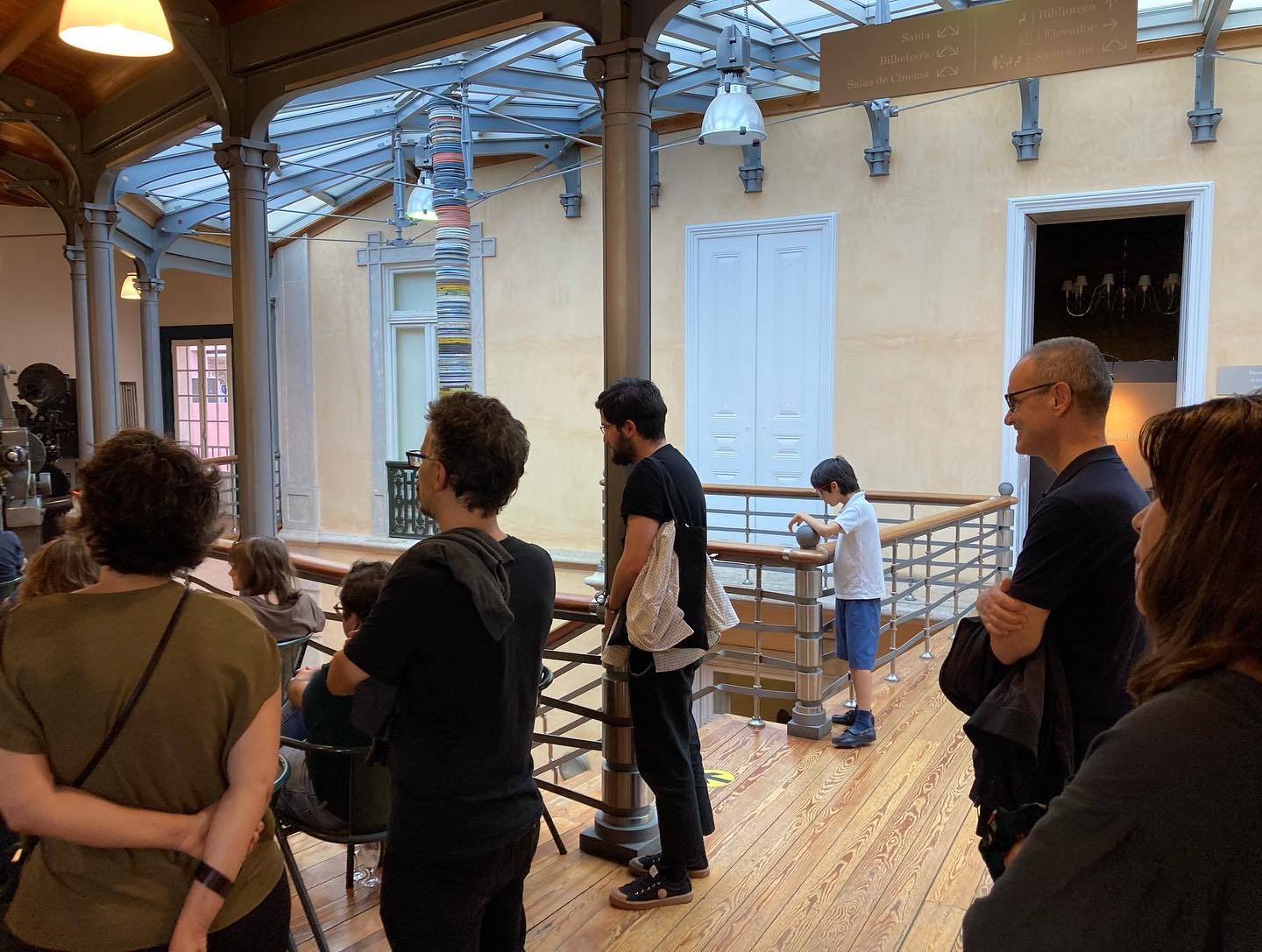 ERVAS ∙ YASUJIRO OZU ∙ JOÃO MIGUEL FERNANDES JORGE ∙ RUI VASCONCELOS ∙ RITA AZEVEDO GOMES Interior com pessoas em pé junto a corrimão de madeira e paredes amarelas.