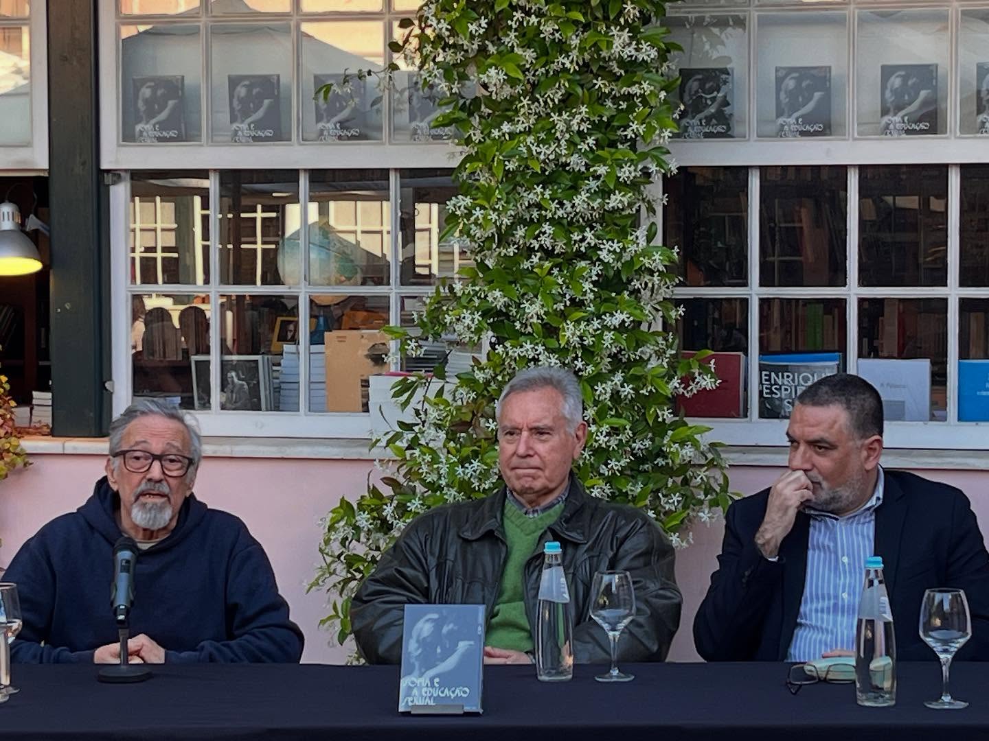 SOFIA E A EDUCAÇÃO SEXUAL ∙ EDUARDO GEADA Homens sentados em mesa preta com livro 'O Fim do Mundo' e garrafas de água em frente a vitrina da livraria