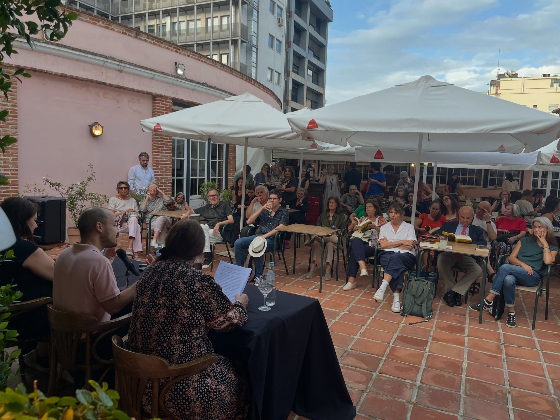 PIONEIRAS DO CINEMA PORTUGUÊS ∙ RICARDO VIEIRA LISBOA (ORG.) Espaço exterior com cadeiras, mesas e guarda-sóis brancos DELTA junto a parede rosa e várias pessoas sentadas.