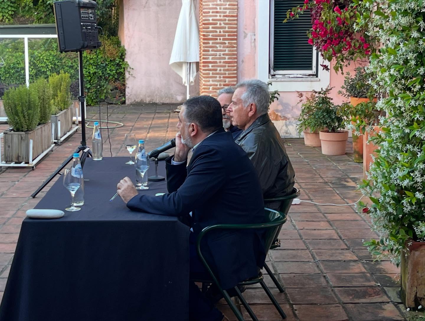 SOFIA E A EDUCAÇÃO SEXUAL ∙ EDUARDO GEADA Mesa preta com copos e garrafas e três homens sentados em ambiente exterior com plantas