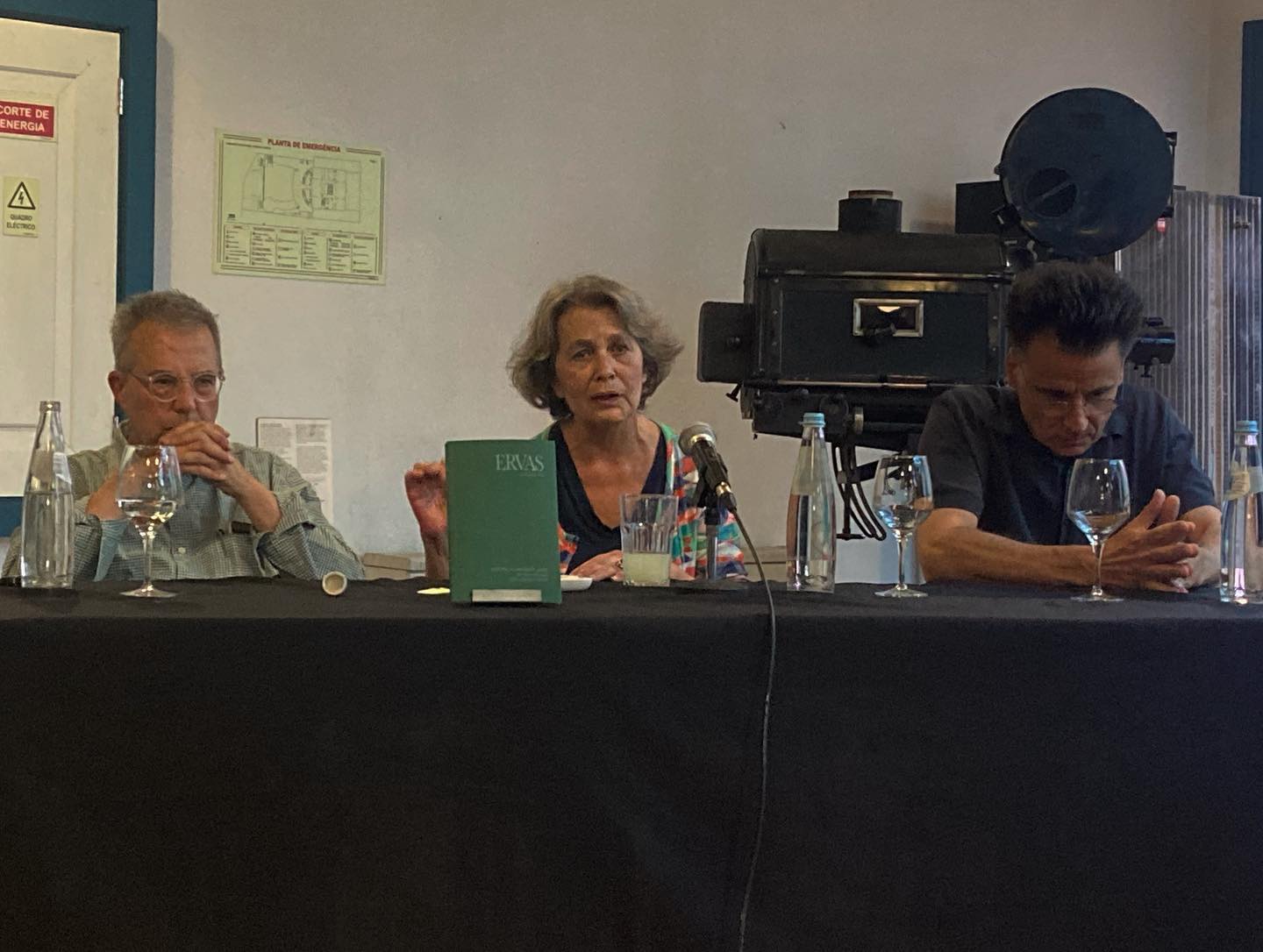 ERVAS ∙ YASUJIRO OZU ∙ JOÃO MIGUEL FERNANDES JORGE ∙ RUI VASCONCELOS ∙ RITA AZEVEDO GOMES Mesa com pessoas e objetos sobre tampo preto com parede branca ao fundo