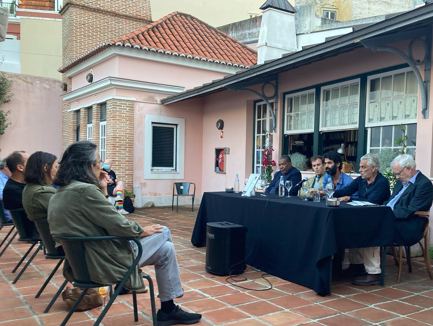 VITALINA VARELA • CADERNO DE RODAGEM • PEDRO COSTA Mesa com toalha preta e copos em pátio com pessoas sentadas em cadeiras pretas