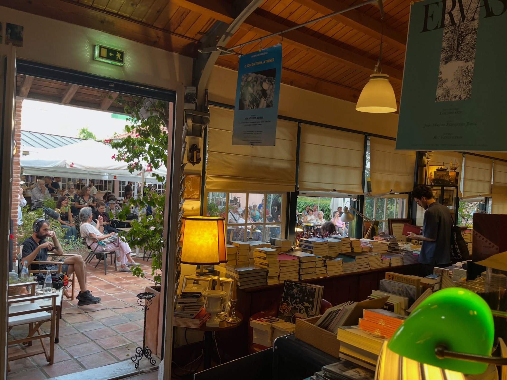 PIONEIRAS DO CINEMA PORTUGUÊS ∙ RICARDO VIEIRA LISBOA (ORG.) Interior de livraria com pilhas de livros, lâmpadas e pessoas sentadas em espaço exterior adjacente