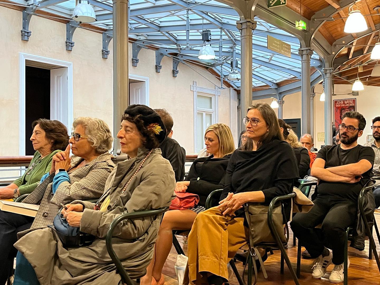 O CINEMA - UMA ESTÉTICA CRUA ∙ JOÃO PAULO DE OLIVEIRA CRUZ MENDES Pessoas sentadas em cadeiras pretas num salão com teto de vidro