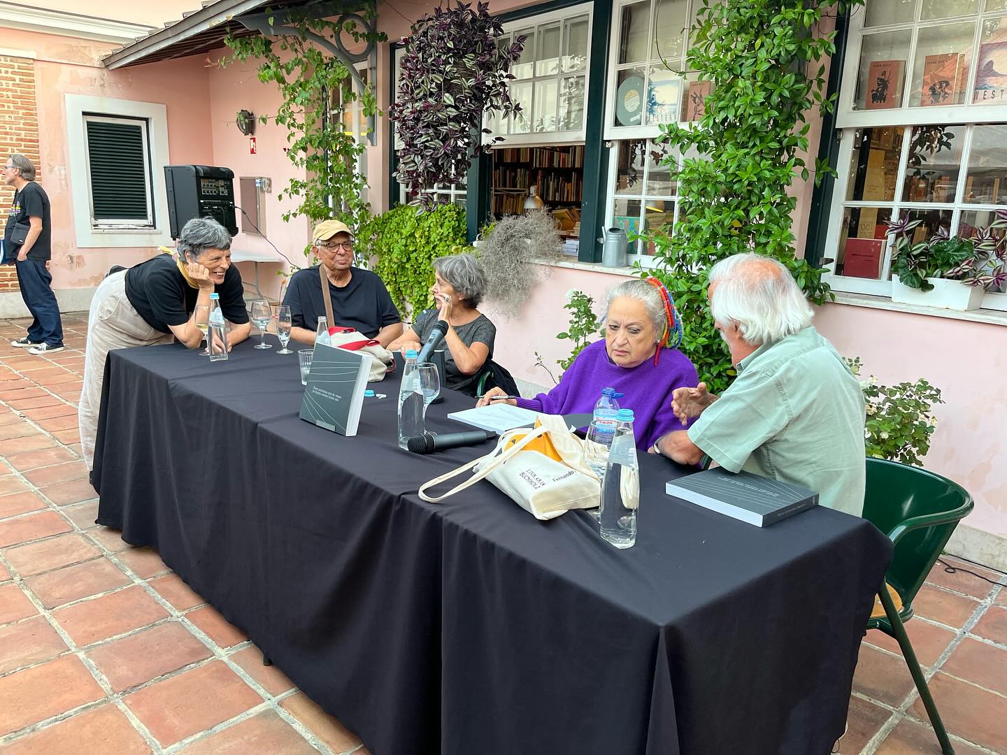 REGINA GUIMARÃES E SAGUENAIL ∙ DUAS PARALELAS QUE ÀS VEZES SE TOCAM OUTRAS VEZES NÃO ∙ INÊS SAPETA DIAS (ORG.) Mesa comprida com toalha preta e pessoas sentadas à volta em espaço exterior com paredes cor de rosa