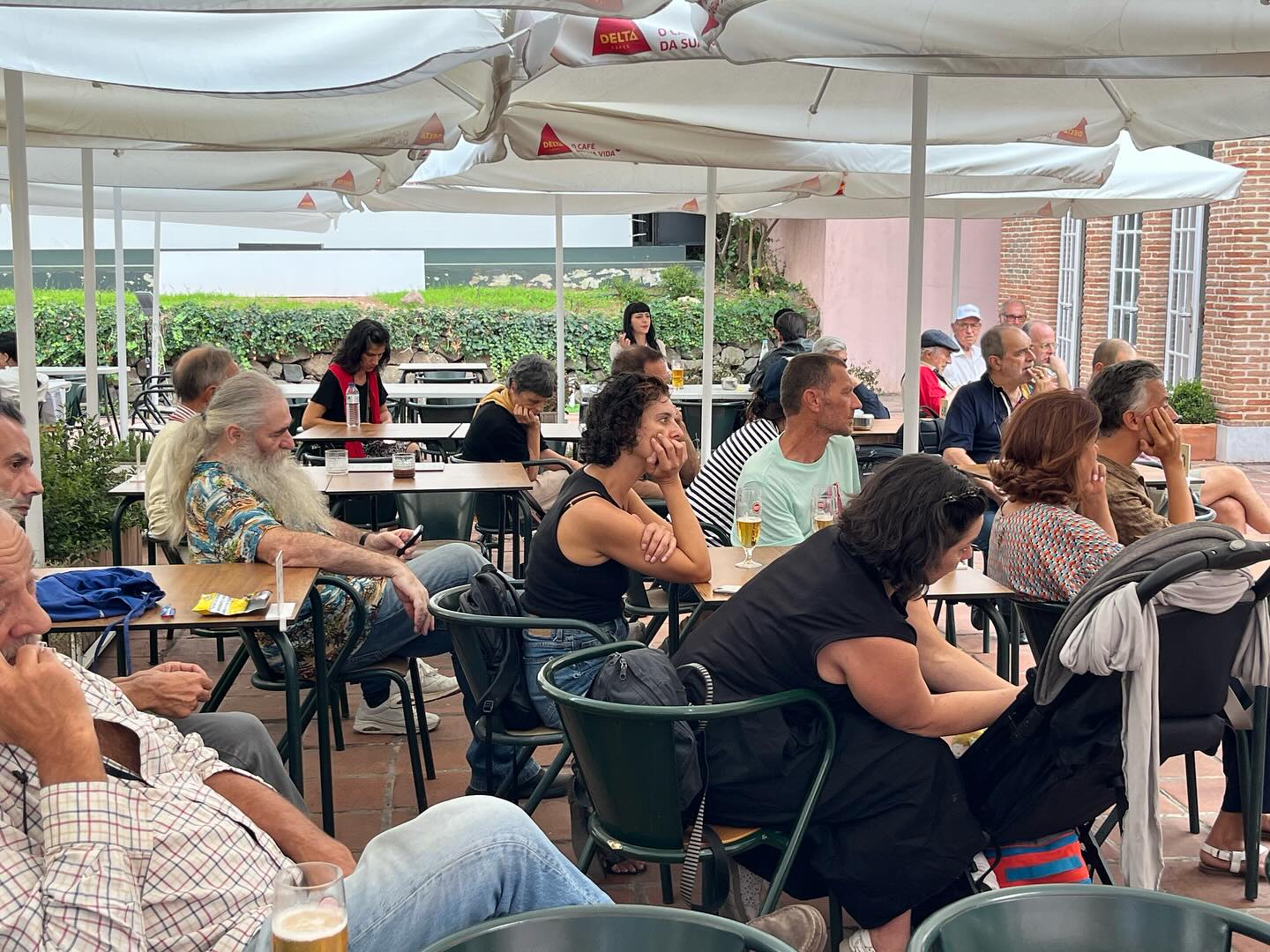 REGINA GUIMARÃES E SAGUENAIL ∙ DUAS PARALELAS QUE ÀS VEZES SE TOCAM OUTRAS VEZES NÃO ∙ INÊS SAPETA DIAS (ORG.) Pessoas sentadas numa esplanada com mesas brancas e cadeiras verdes sob guarda-sóis brancos