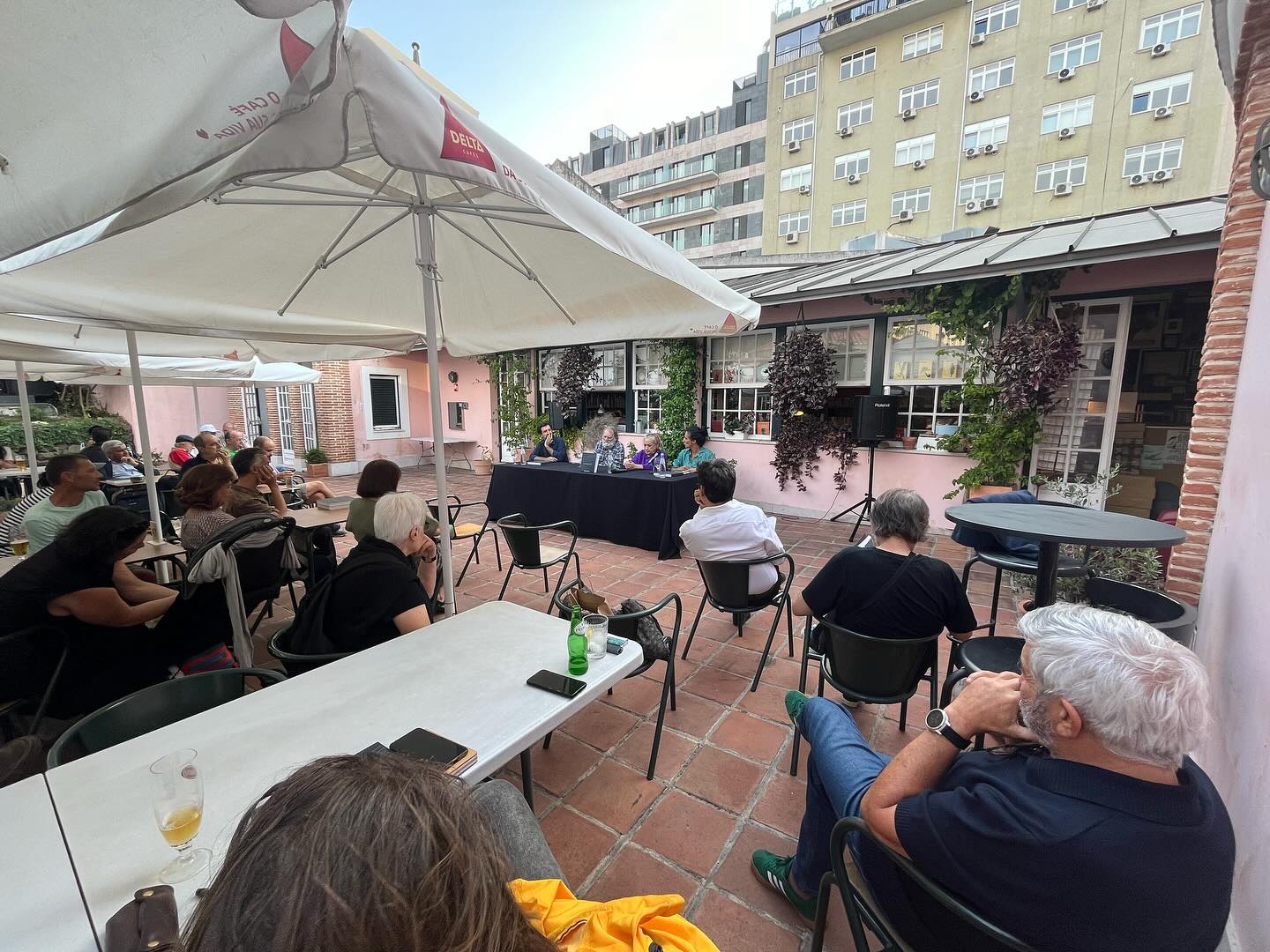 REGINA GUIMARÃES E SAGUENAIL ∙ DUAS PARALELAS QUE ÀS VEZES SE TOCAM OUTRAS VEZES NÃO ∙ INÊS SAPETA DIAS (ORG.) Pessoas sentadas em cadeiras num espaço exterior com mesas e guarda-sóis brancos junto a edifícios.