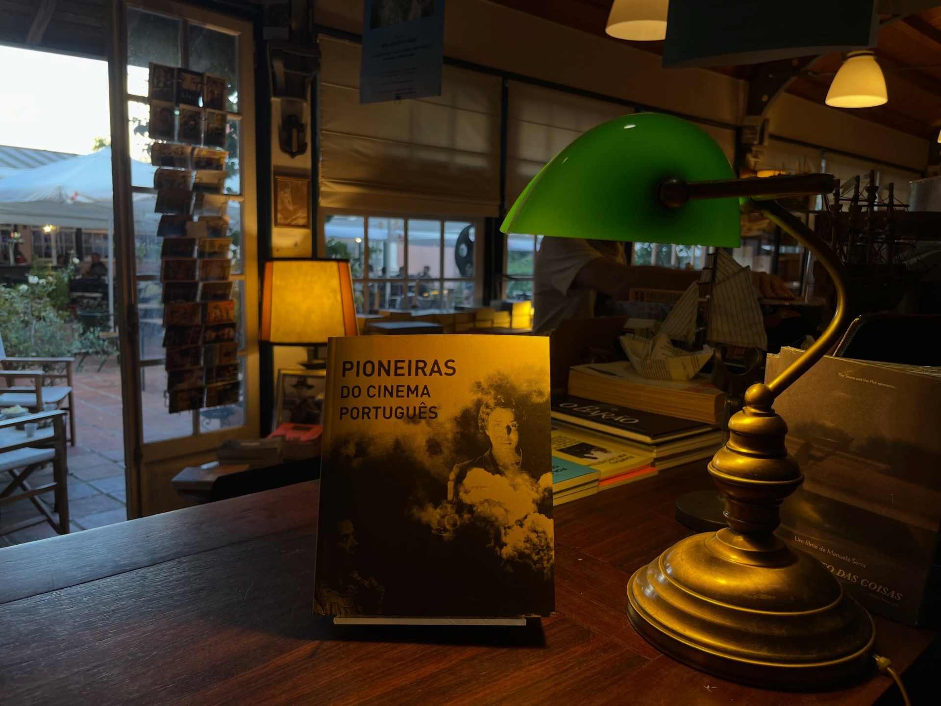 PIONEIRAS DO CINEMA PORTUGUÊS ∙ RICARDO VIEIRA LISBOA (ORG.) Livro PIONEIRAS DO CINEMA PORTUGUÊS sobre mesa de madeira ao lado de candeeiro verde numa sala com janelas e iluminação quente