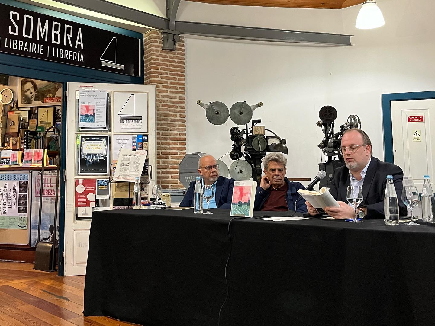 O CINEMA - UMA ESTÉTICA CRUA ∙ JOÃO PAULO DE OLIVEIRA CRUZ MENDES Três homens numa mesa de apresentação com livros e garrafas de água, estante de livros e equipamentos vintage ao fundo
