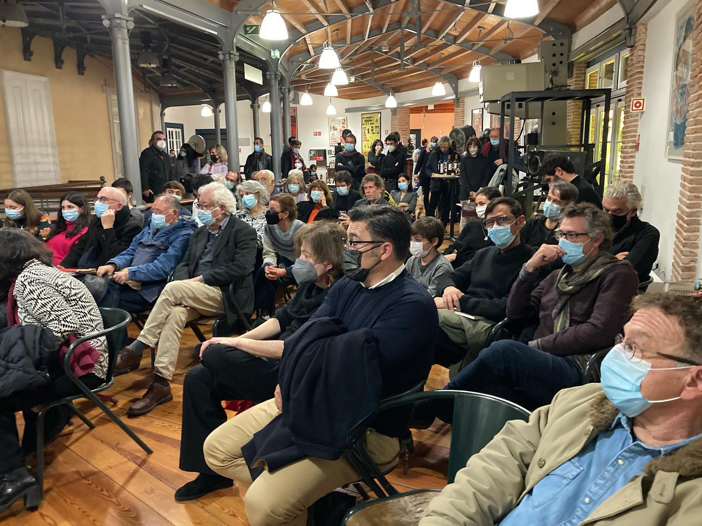 DIZER O MUNDO • CONVERSAS COM RUI NUNES E PAULO NOZOLINO • ALEXANDRA CARITA Pessoas sentadas em cadeiras num espaço interior com teto de madeira e paredes de tijolo